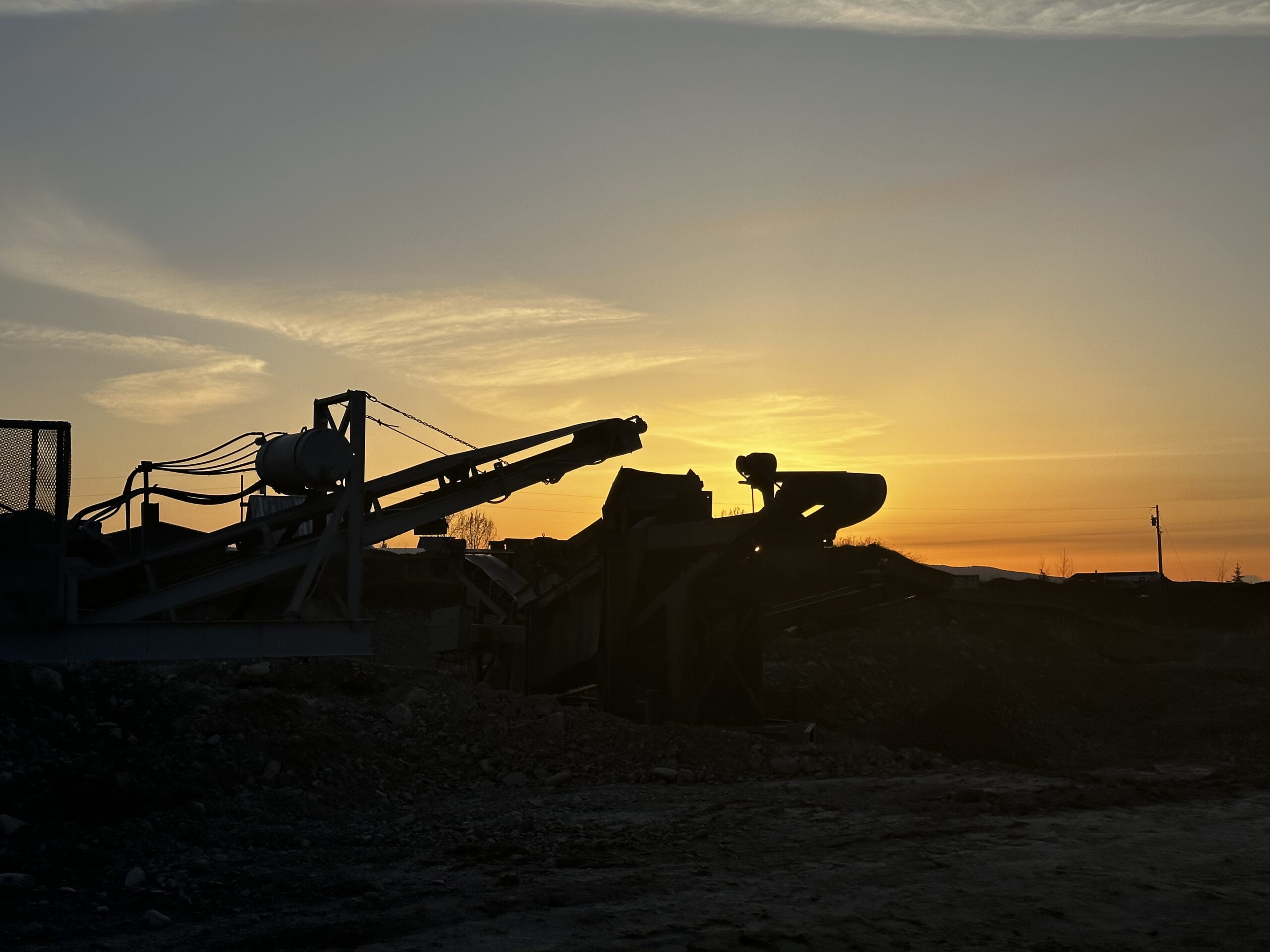 Darby Pit, Gravel & Trucking - We specialize in supplying top-quality materials such as dirt, sand, gravel, crushed stone, and providing reliable trucking services for all your needs. Whether you're working on excavation projects, landscaping endeavors, home construction, road building, paving paths and driveways, or laying down foundations – we've got you covered. Against the backdrop of a golden sunset sky with the silhouette of mining machinery outlined against rugged terrain and faint distant buildings – we ensure that every project is supported with the best materials and services in the industry.