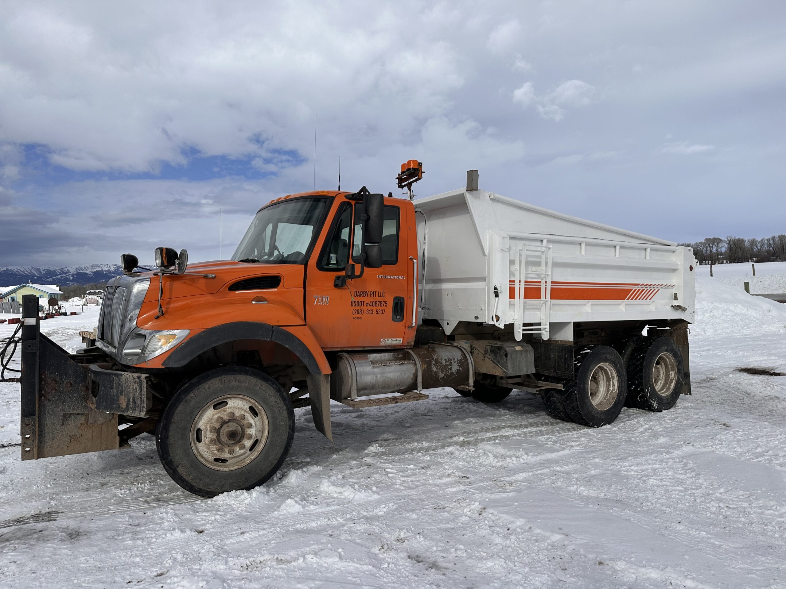 Darby Pit, Gravel & Trucking - We specialize in supplying a comprehensive range of materials including dirt, sand, gravel, and crushed stone. Our services cover trucking for excavation projects, landscaping needs, home building endeavors, and constructing roads, paths, driveways, and foundations. Featured in our fleet is an orange and white snowplow truck with a snowblade ready for action. It stands poised on a snowy ground under a cloudy sky, illustrating our capability to handle winter conditions with efficiency.