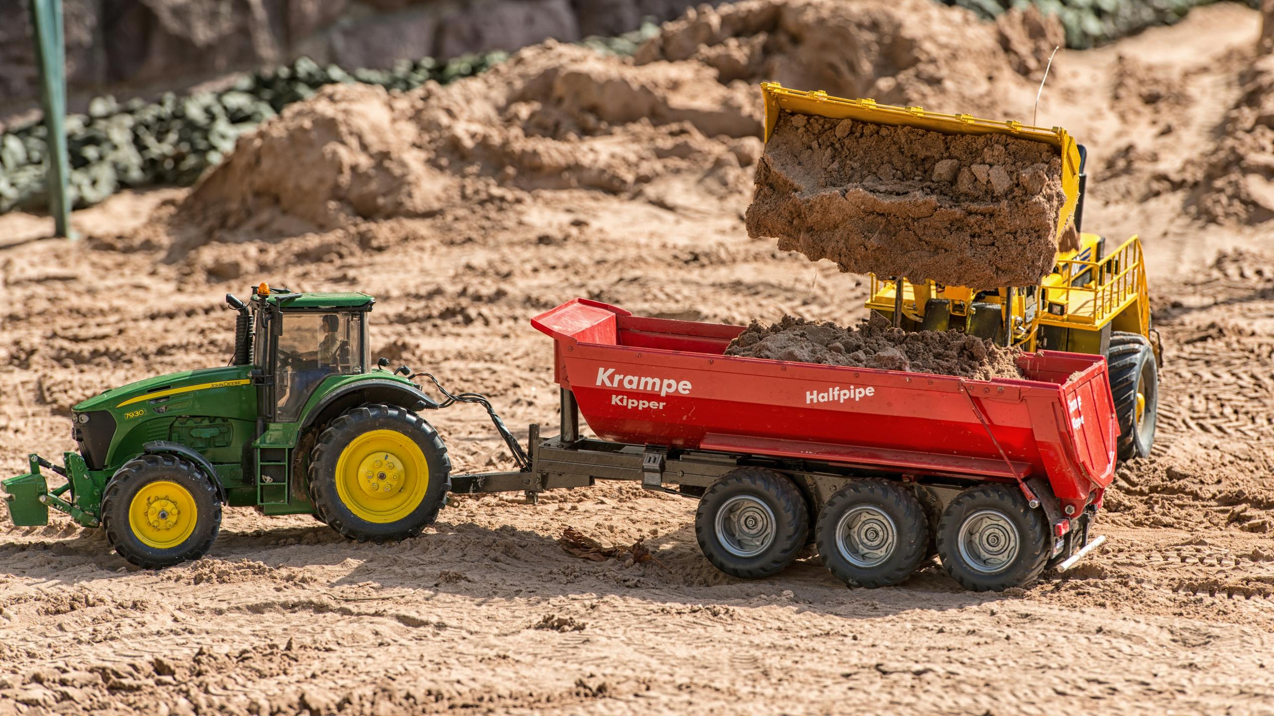 Close-up of toy excavator loading soil into a tractor trailer on a sandy landscape.