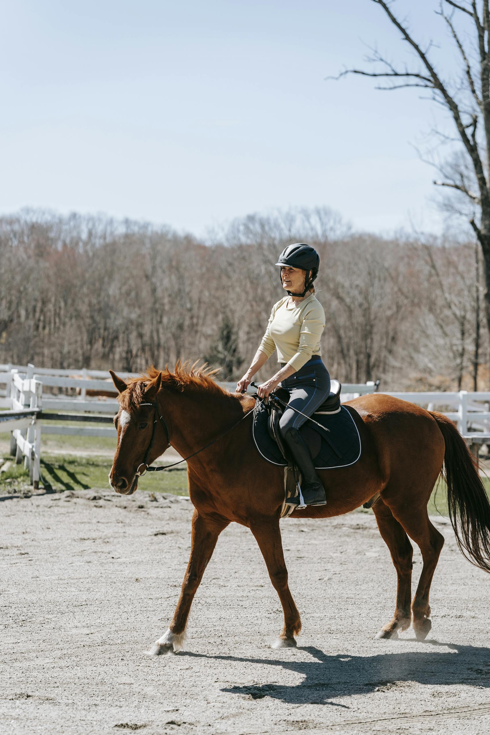 Young woman horseback riding in a sunny outdoor arena, wearing a helmet and casual attire.